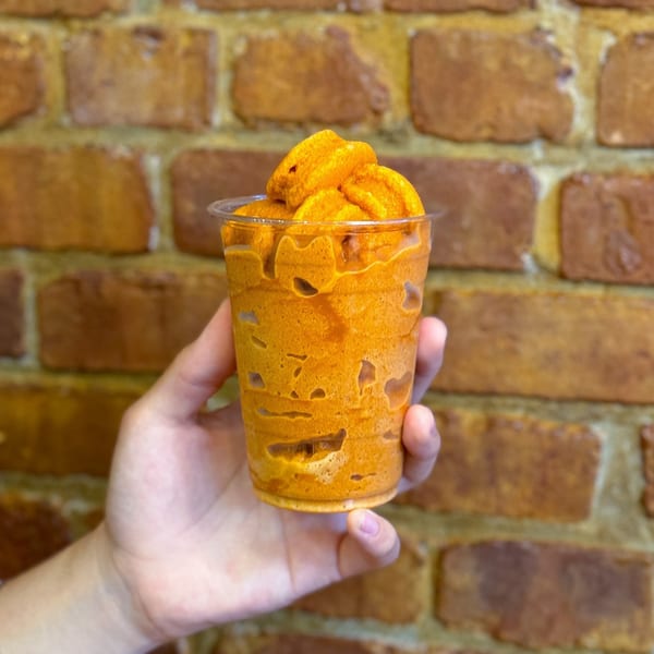 A hand holds a Thai iced tea slushie with a brick wall background at Gai Kitchen.