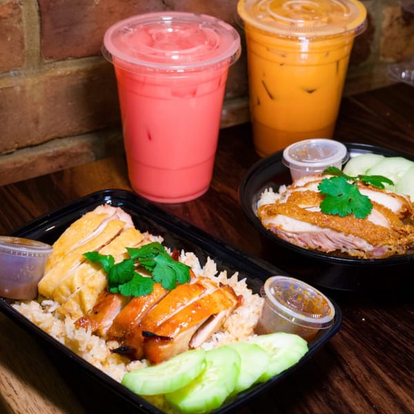 Two Thai chicken and rice dishes with cucumbers, sauces, and two iced drinks on a wooden table.