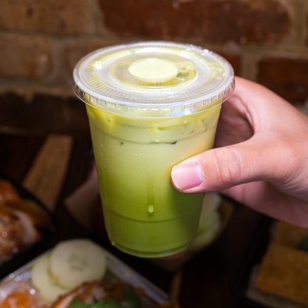 Hand holding a plastic cup of iced green Thai tea with condensation, against a brick wall.