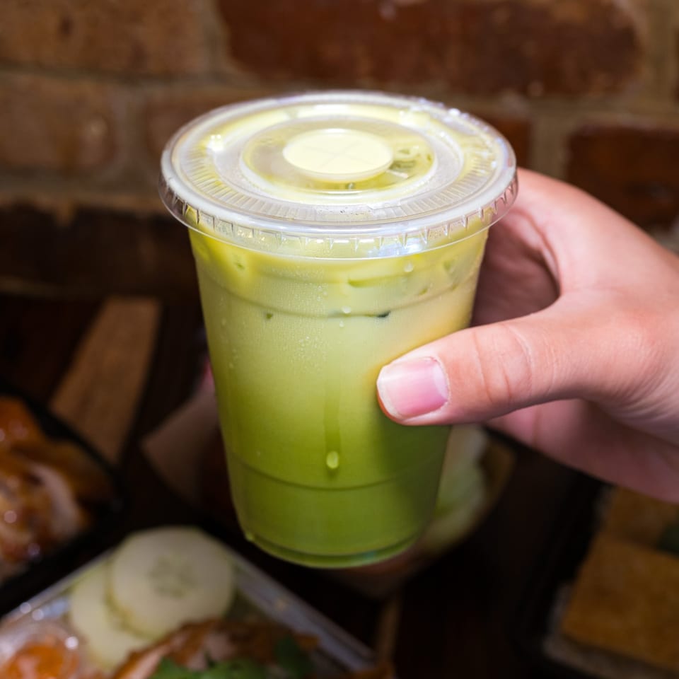 Hand holding a plastic cup of iced green Thai tea with condensation, against a brick wall.