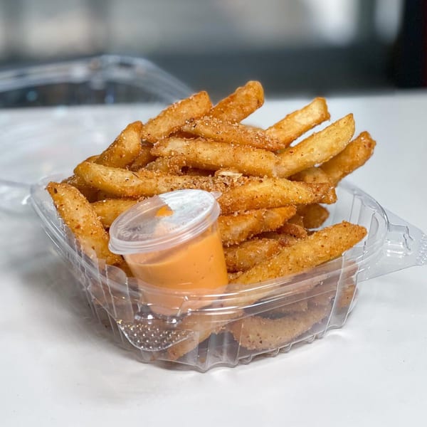 Golden brown french fries seasoned with spices, served in a clear plastic container with dipping sauce.