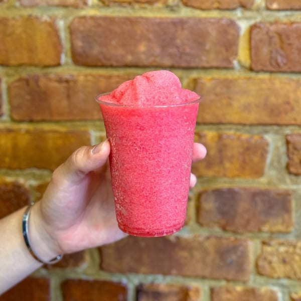 Hand holding a vibrant pink Thai iced slushy drink against a rustic brick wall.