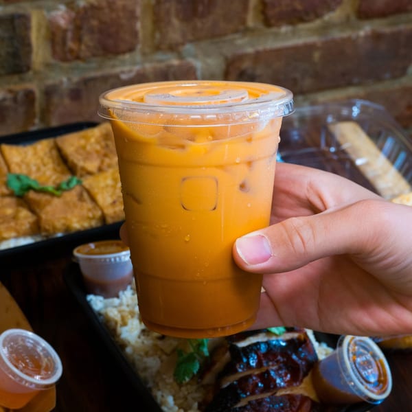A hand holds a plastic cup of iced Thai tea, with food containers blurred in the background.