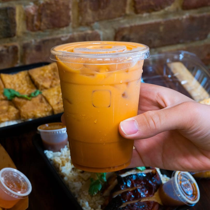 A hand holds a plastic cup of iced Thai tea, with food containers blurred in the background.