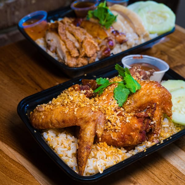 Two Gai Kitchen Thai chicken and rice dishes in black takeout containers on a wooden table.