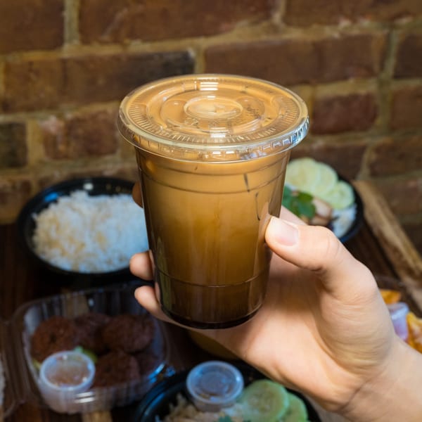 Hand holding a Thai iced tea in a plastic cup, with other Thai dishes blurred in the background.