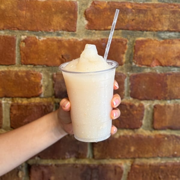 Hand holding a plastic cup of light-colored Thai iced tea slushie with a straw against a brick wall.